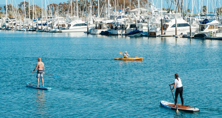 A man in a yellow kayak paddling opposite a woman and a man standing on their SUP-boards near a long pier with white water crafts