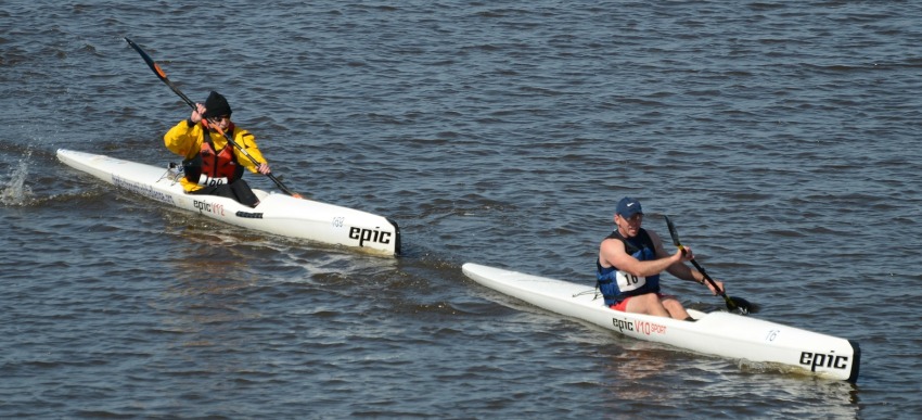Two men with numbers on their chests paddling long white kayaks during the race