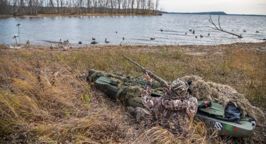 A hunter sits in an ambush along with his kayak, aiming his rifle at some target in the air 