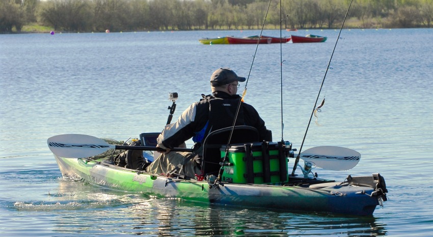A fisherman, surrounded by fishing rods and accessories, paddles his green kayak on the water