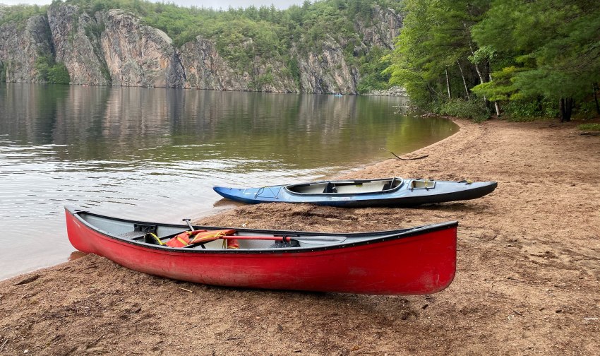 A red canoe and a blue kayak lie on the shore close to some picturesque cliffs