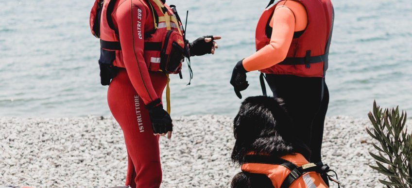 Two people and a dog wearing red life jackets on the beach