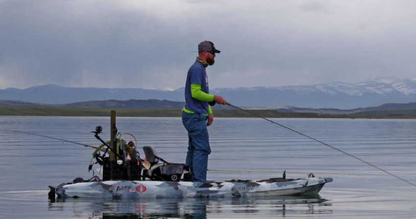 A man with a fishing rod standing on a kayak, loaded with other fishing gear