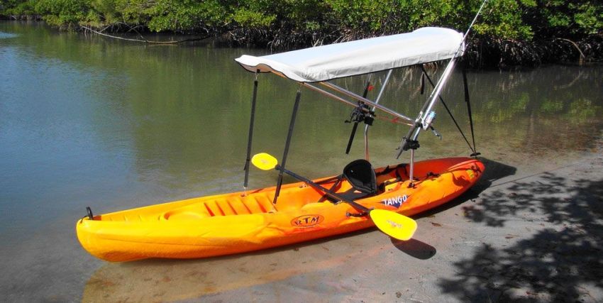 An orange kayak with a bimini, a paddle  and two fishing rods lies on the river bank