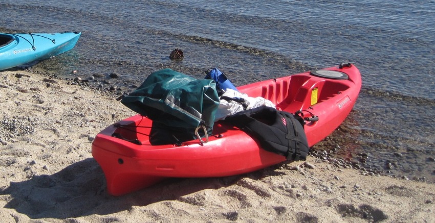 A red kayak with a green dry bag and human clothes lies on the shore