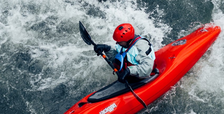 A man in grey drysuit and red helmet paddles his red kayak in the whitewater