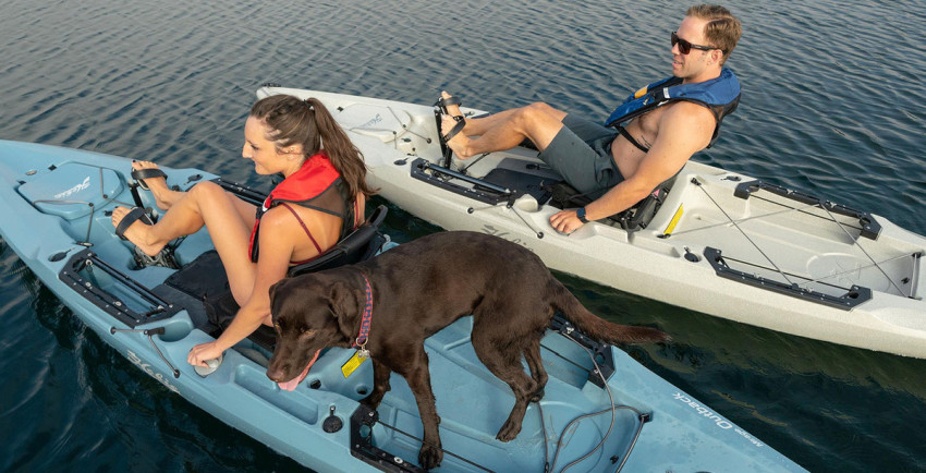 A young lady with her dog and a young man happily pedal their kayaks on the water
