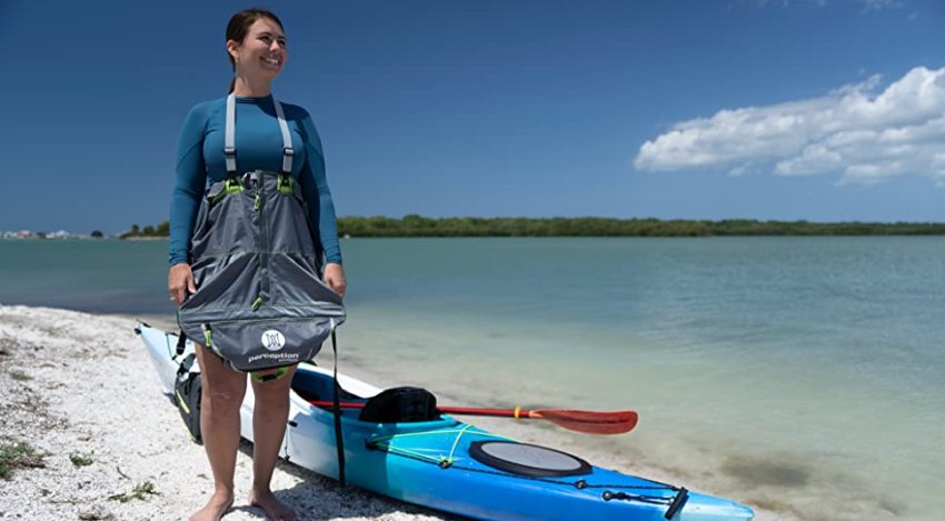 A woman wearing a spray skirt stands new her blue kayak on the shore