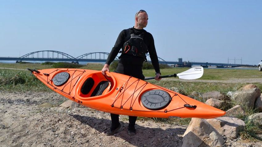 A man carries an orange sit-in kayak and a paddle