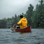 A man wearing a yellow coat paddles his red canoe on the water