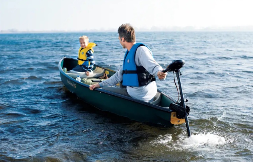 A man sits in a green canoe and controls a trolling motor, while looking at his kid in a yellow PFD, sitting on the bow.