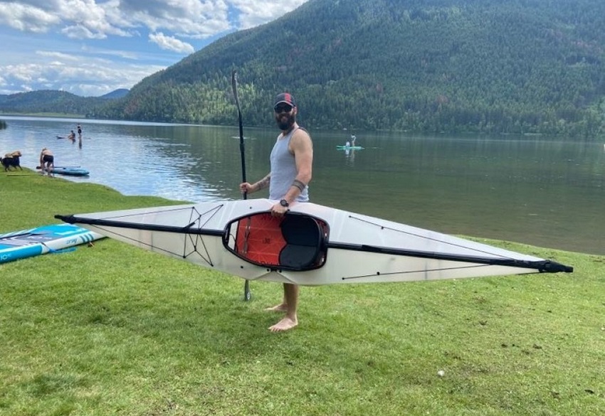 A man stands on a lake shore and holds a white Oru kayak and a black paddle 