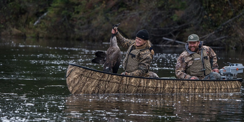 A female hunter sits in the Esquif Mallard canoe, holding a duck in her hand. A male hunter behind her controls the canoe's motor.