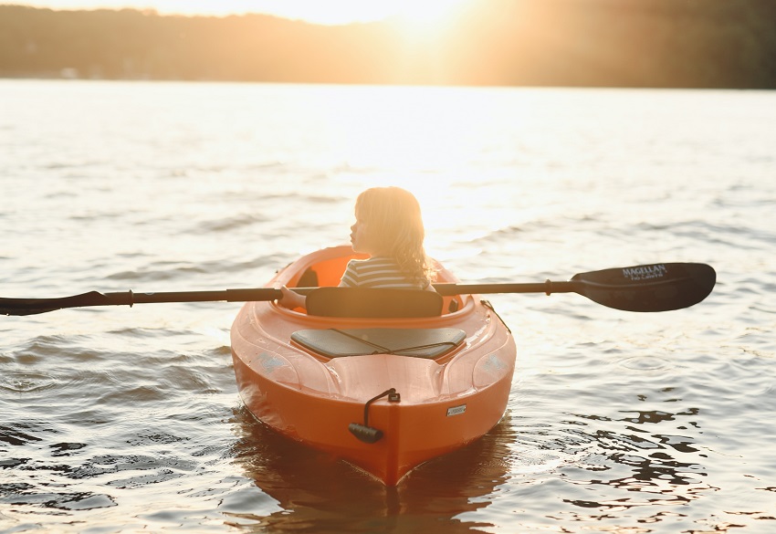 A kid with a black paddle in his hands sits in an red kayak, facing the sunset
