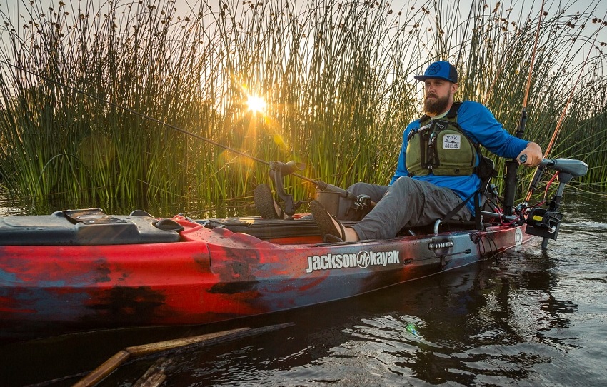 An angler sits on a red and blue Jackson kayak and controls a trolling motor