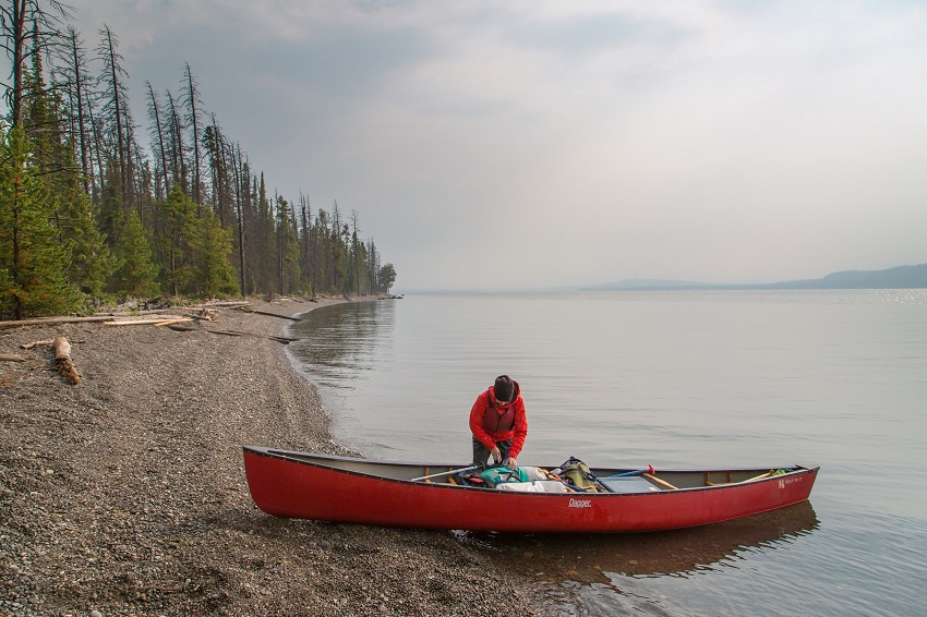 A paddler, wearing a red weatherproof jacket, packs camping gear in a red canoe on a shingle beach