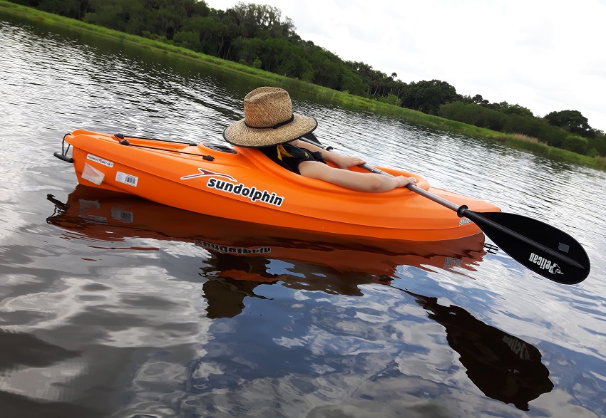 A woman with a black paddle in her hands relaxes in an orange recreational kayak on water 