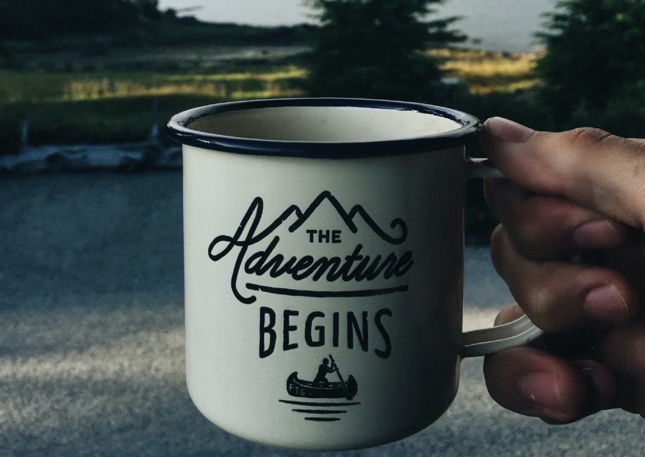 A human hand holds a white mug with "The Adventure Begins" caption
