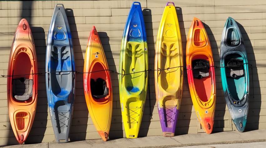 Seven plastic kayaks of different colors stand leaned against a wall
