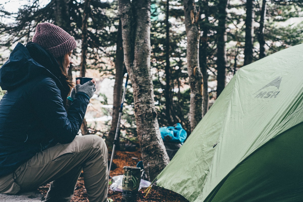 A woman with a cup of coffee sits in front of a tent in a forest