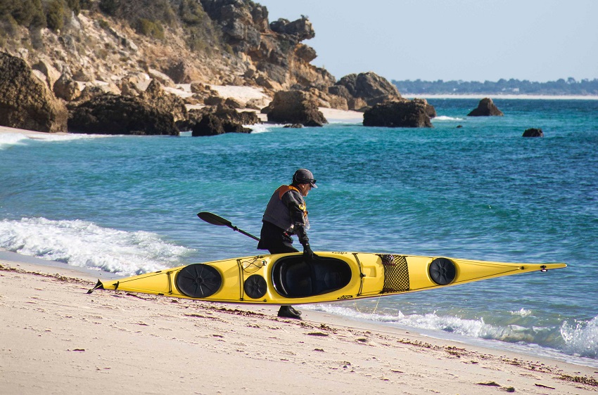 A paddler carries his yellow sit-in kayak on the beach towards the water