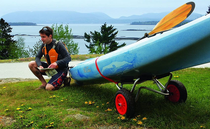 A man, holding a map in his hands, is sitting near a blue kayak on a kayak trolley.