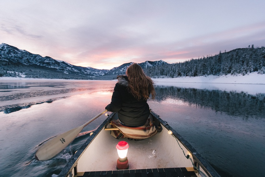 A woman with a paddle in hands sits at the canoe bow on the lake, surrounded by snowy forests and mountains