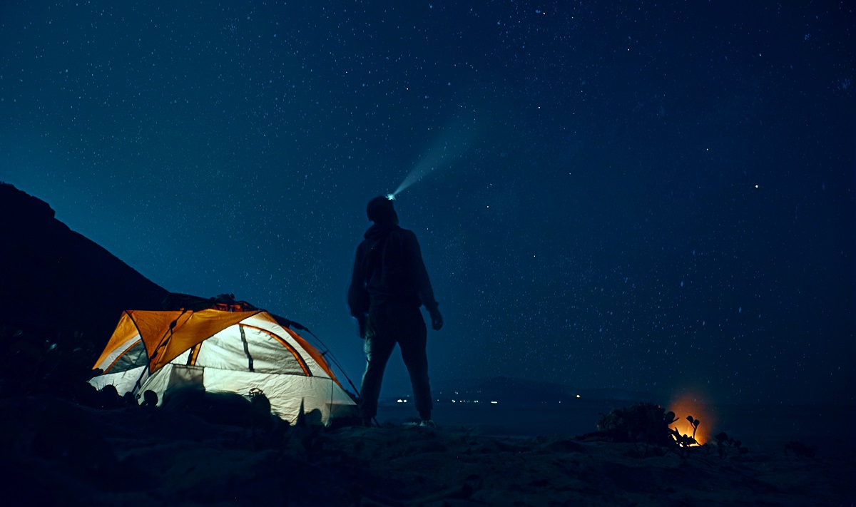 A man with a headlight on his head stands near the lighted tent and observes the night sky