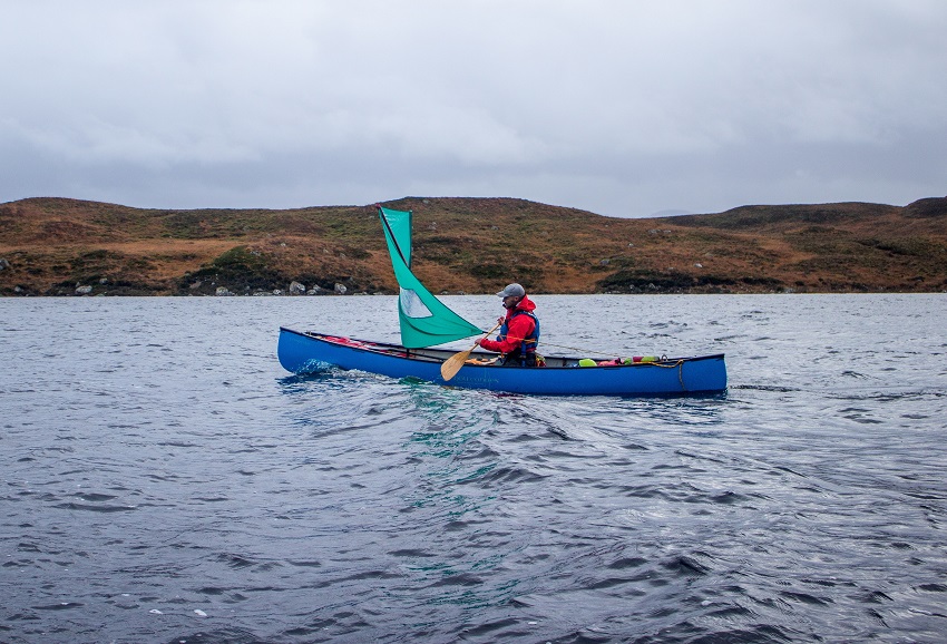A man paddles a blue canoe with a green sail