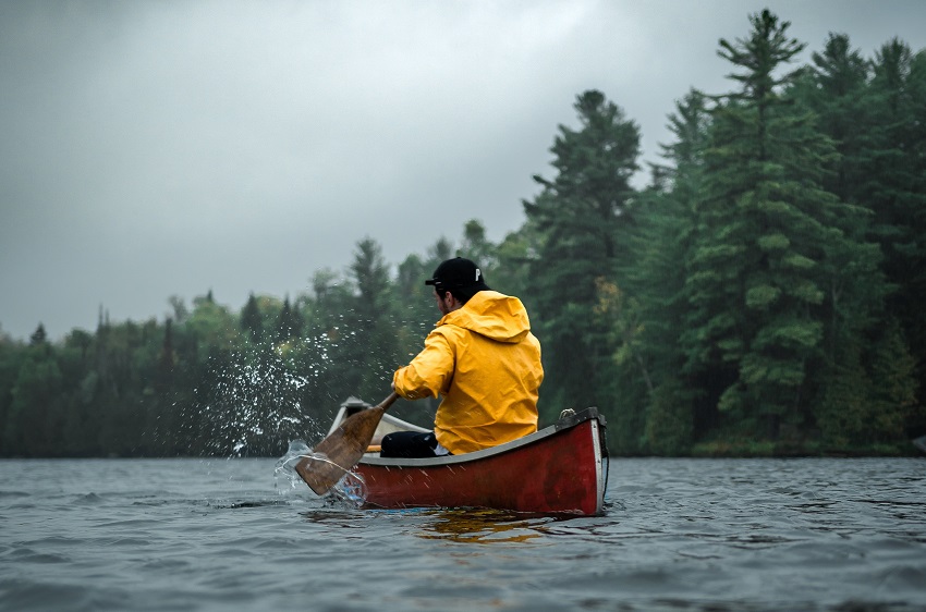 A man in a yellow raincoat paddles a red solo canoe along the woody coast