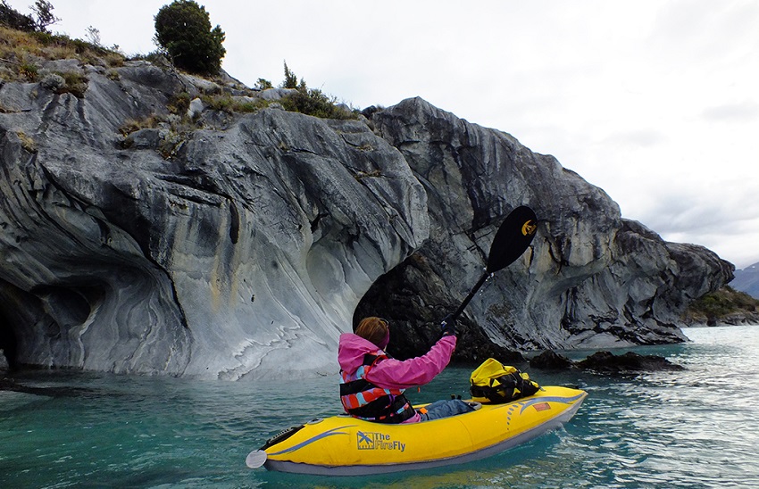 A woman in a pink jacket and a red PFD paddles a yellow Advanced Elements Firefly kayak along the cliffs