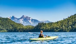 A woman paddles a green kayak on open water surrounded by trees and mountains
