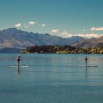 Two women paddle their boards on a lake surrounded by mountains