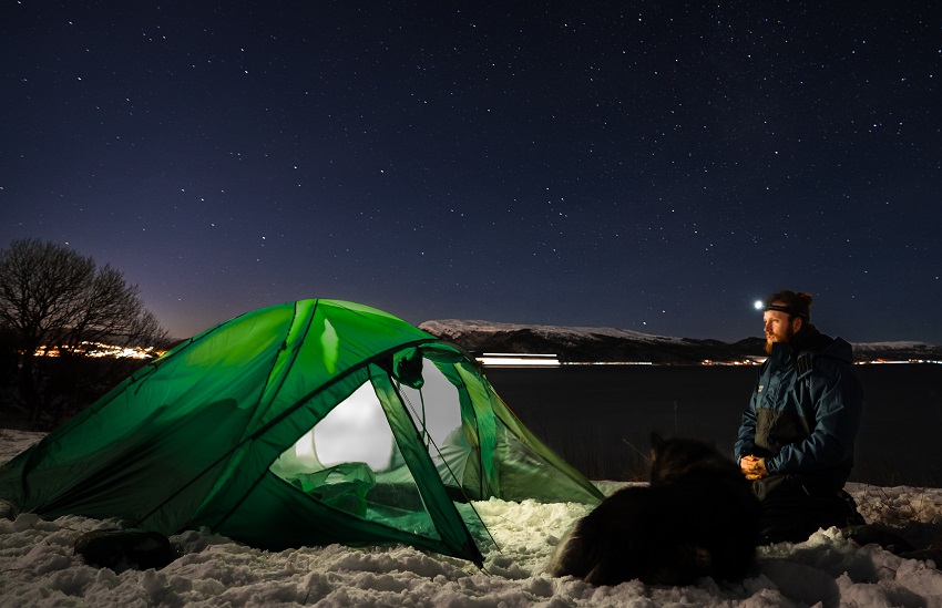 A man with a headlight and a dog sit in front of a green tent at night