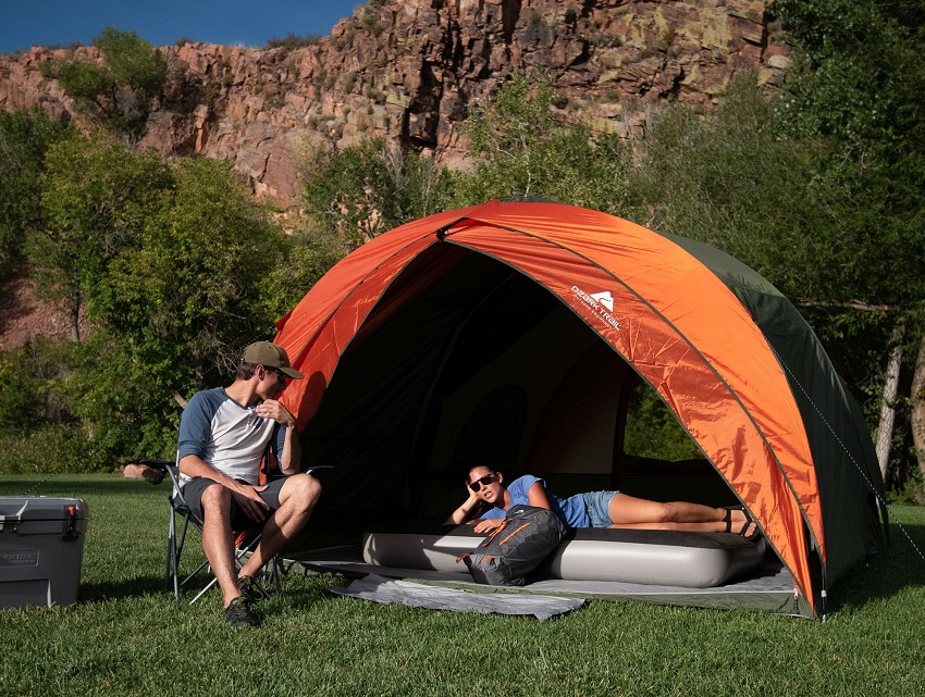A man sits in a chair and talks to a woman lying on an air mattress in the Ozark Trail 8-Person Dome Tunnel Tent, with Maximum Weather Protection