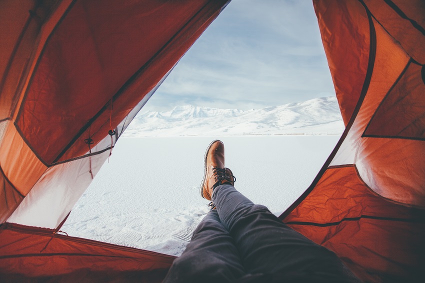 Human legs are stretched out of a camping tent towards the snowy mountain view