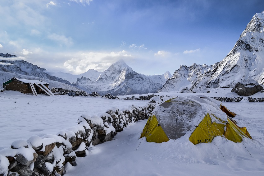 A yellow tent is set up near a stone wall, surrounded by snowy mountains