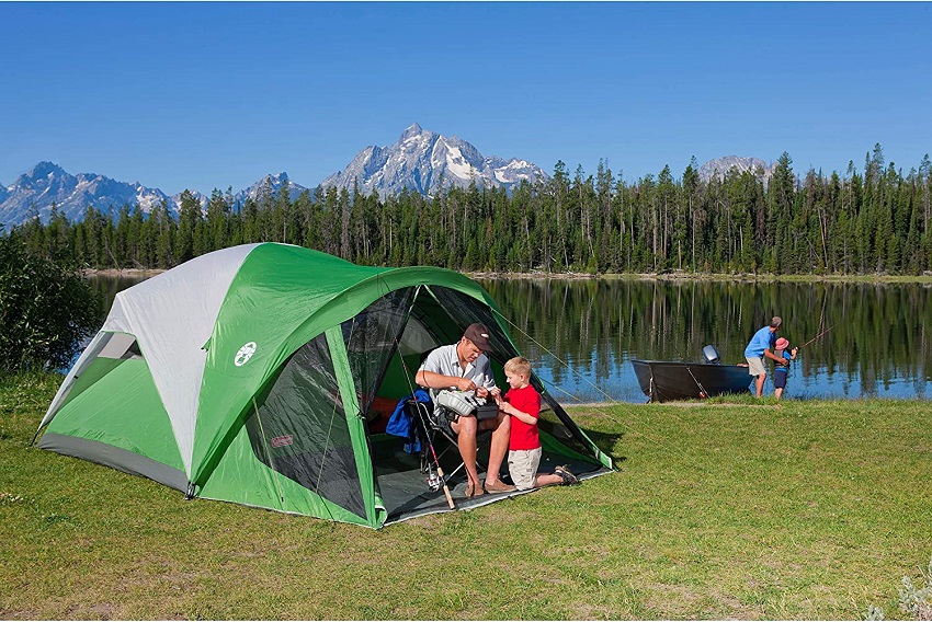 A man and a child prepare their fishing gear inside a green and white camping tent. Another man helps his kid to cast a line in the nearby lake.