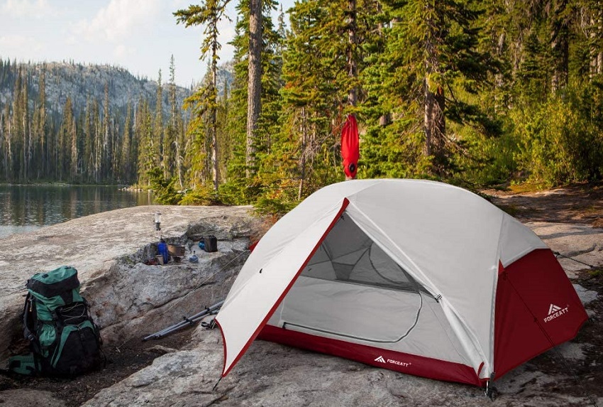 A white and red camping tent, a green backpack and camping cookware on a rocky lake shore