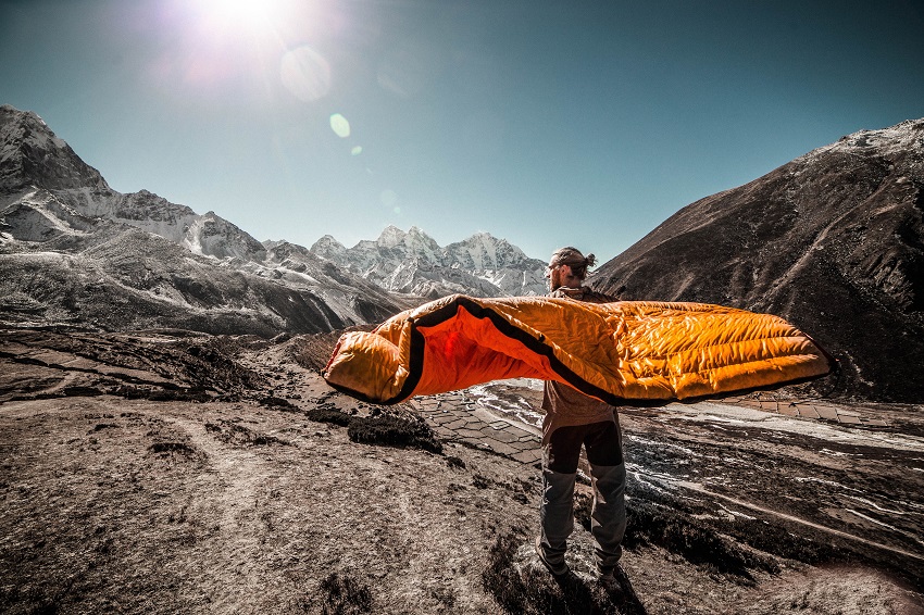 A man stands on a rocky trail way and holds an orange sleeping bag on his shoulders