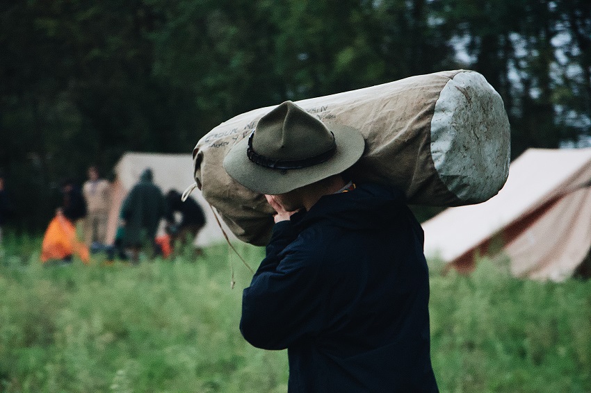 A man in a green hat carries a packed tent on his shoulder