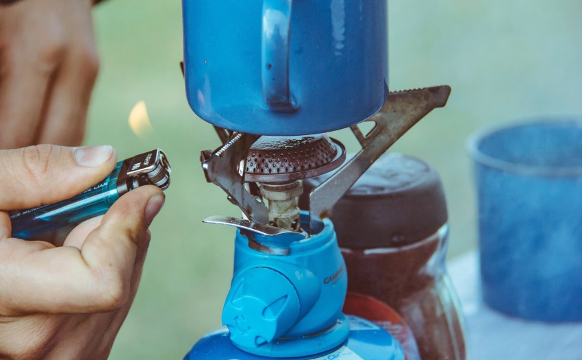 A human hand lights a camping gas stove, bearing a blue cup, with a lighter 