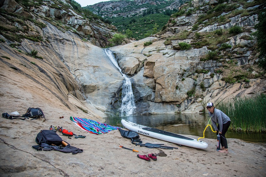 A woman inflates a white inflatable paddle board on the stony shore near a waterfall