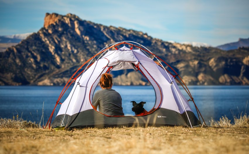 A woman and her black dog sit inside a camping tent, pitched near a lake