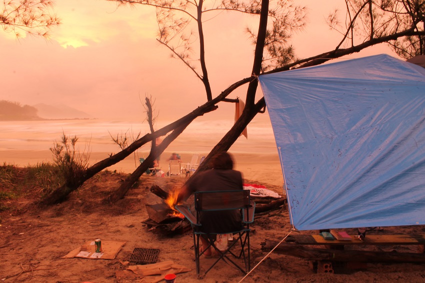 A man sits in a camping chair near the campfire under a blue tarp, stretched between trees