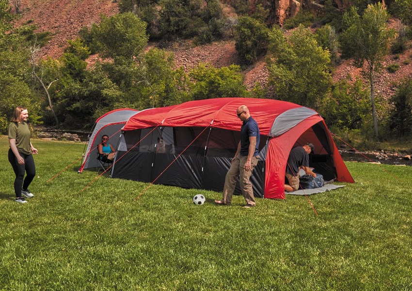 A man and a woman play football in front of the red Ozark Trail 10-Person Tunnel Tent. Another woman sits in a chair in that tent's vestibule.