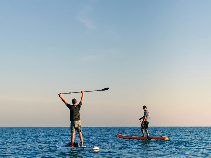 One man paddles an orange SUP, another man stands on his SUP nearby hands up with a paddle