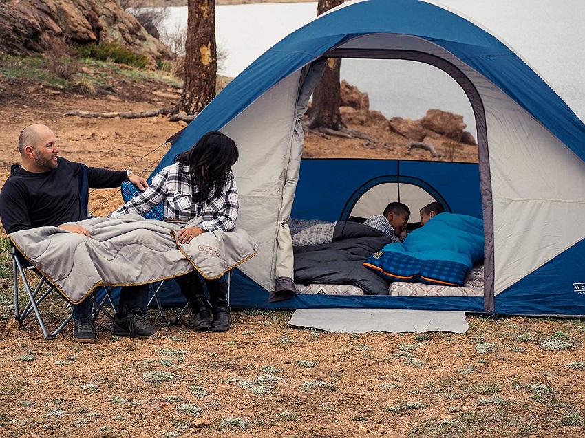 A man and a woman sit in camping chairs and look at their children, lying in sleeping bags inside the Wenzel Tamarack 6-Person Dome tent