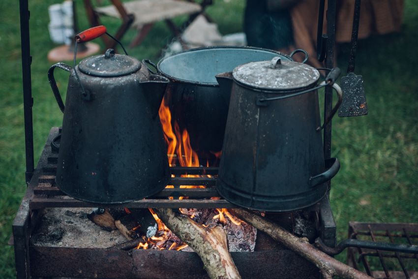 A pan and two teapots are heated on a grille over a campfire