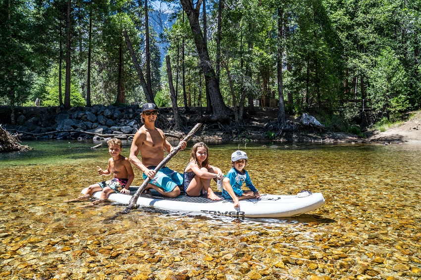 A man with three kids paddle their SUP on a shallow river